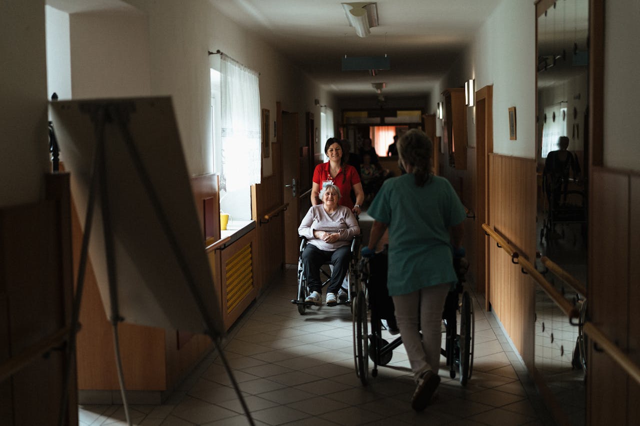Caregiver assisting elderly woman in a wheelchair through a retirement home corridor.